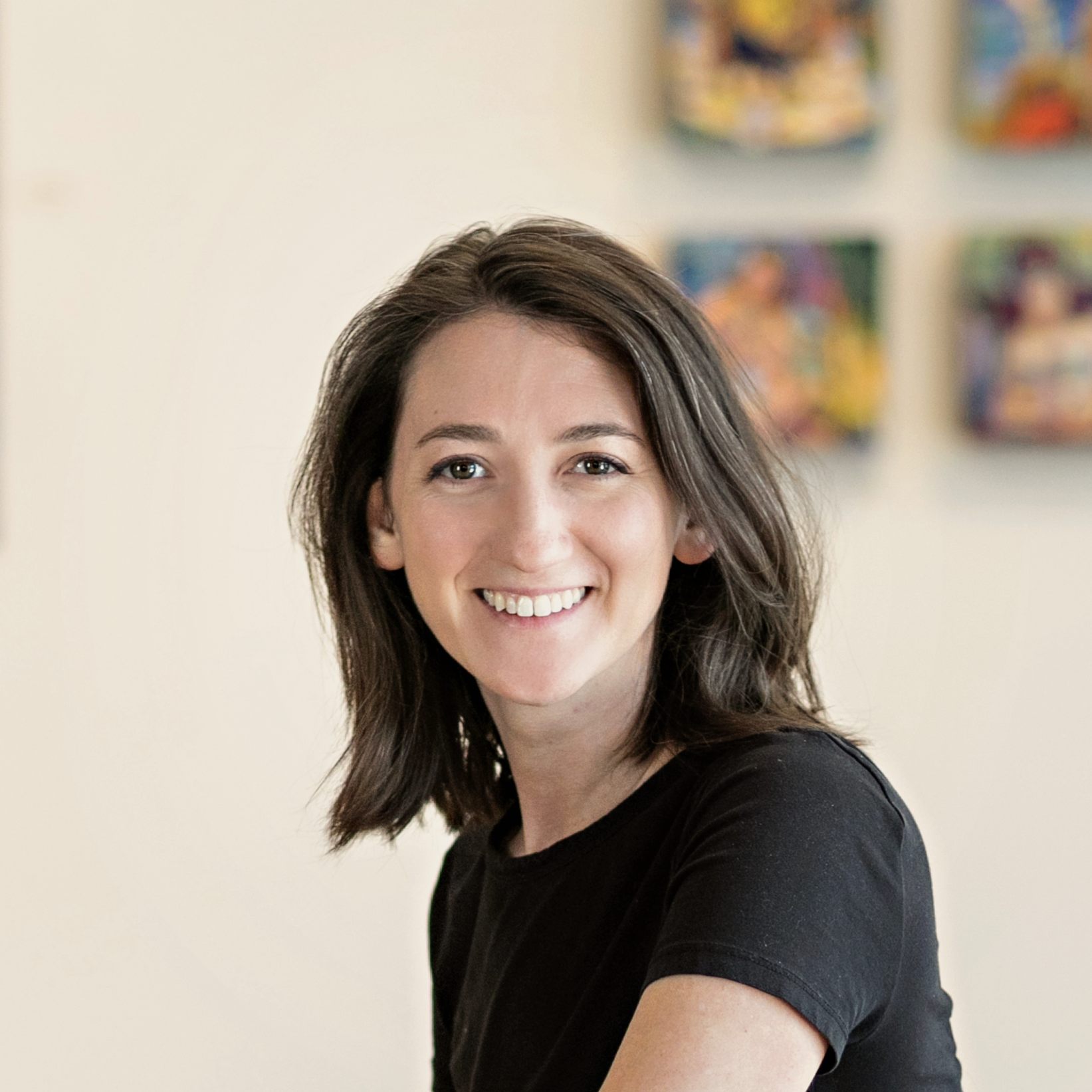 A smiling woman with shoulder-length brown hair wearing a black short-sleeve top, photographed indoors against a light beige wall with colorful framed art blurred in the background.