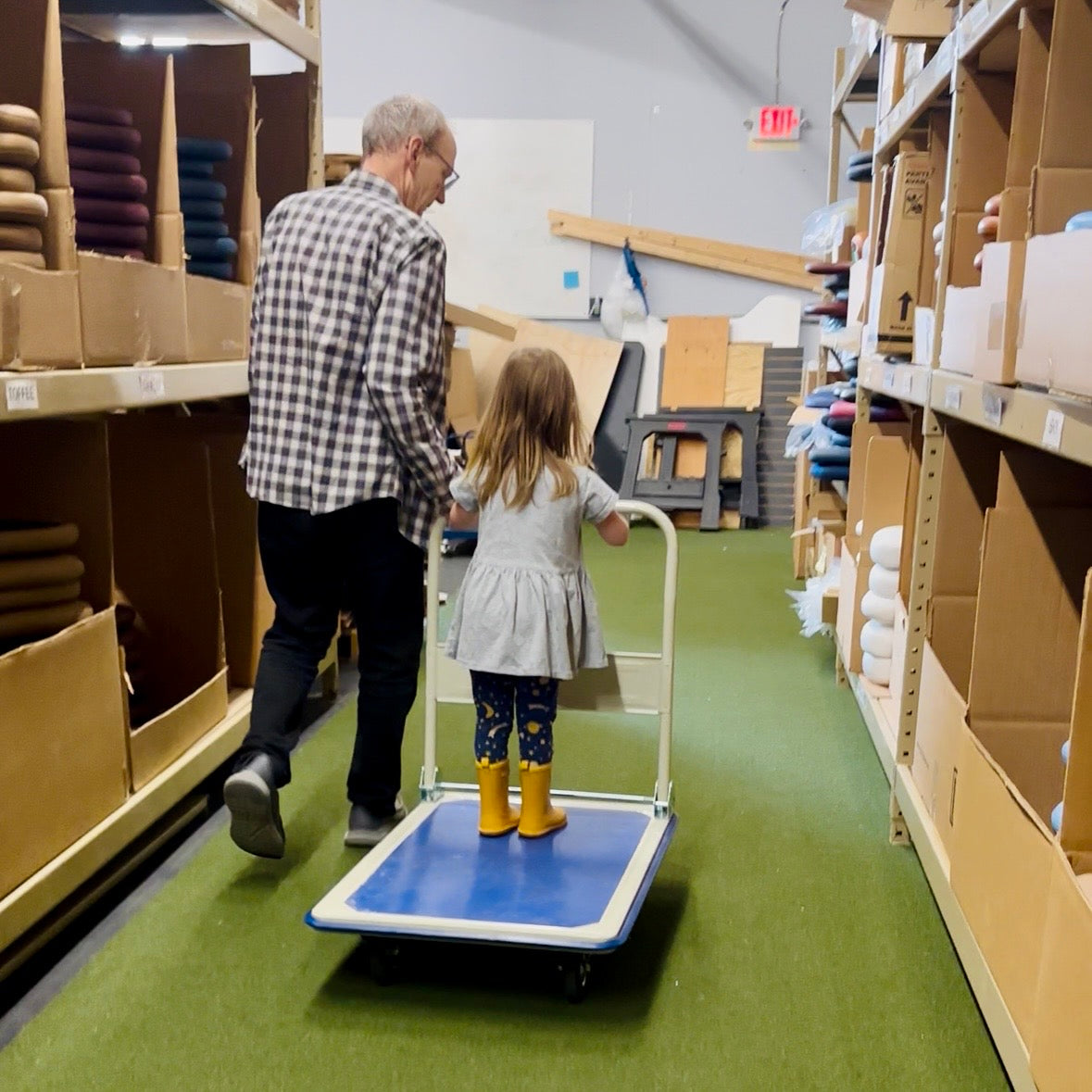 A man and a young girl push a blue and white hand truck through an aisle in a warehouse or retail storage area with shelves of boxes and soul seat cushions on both sides