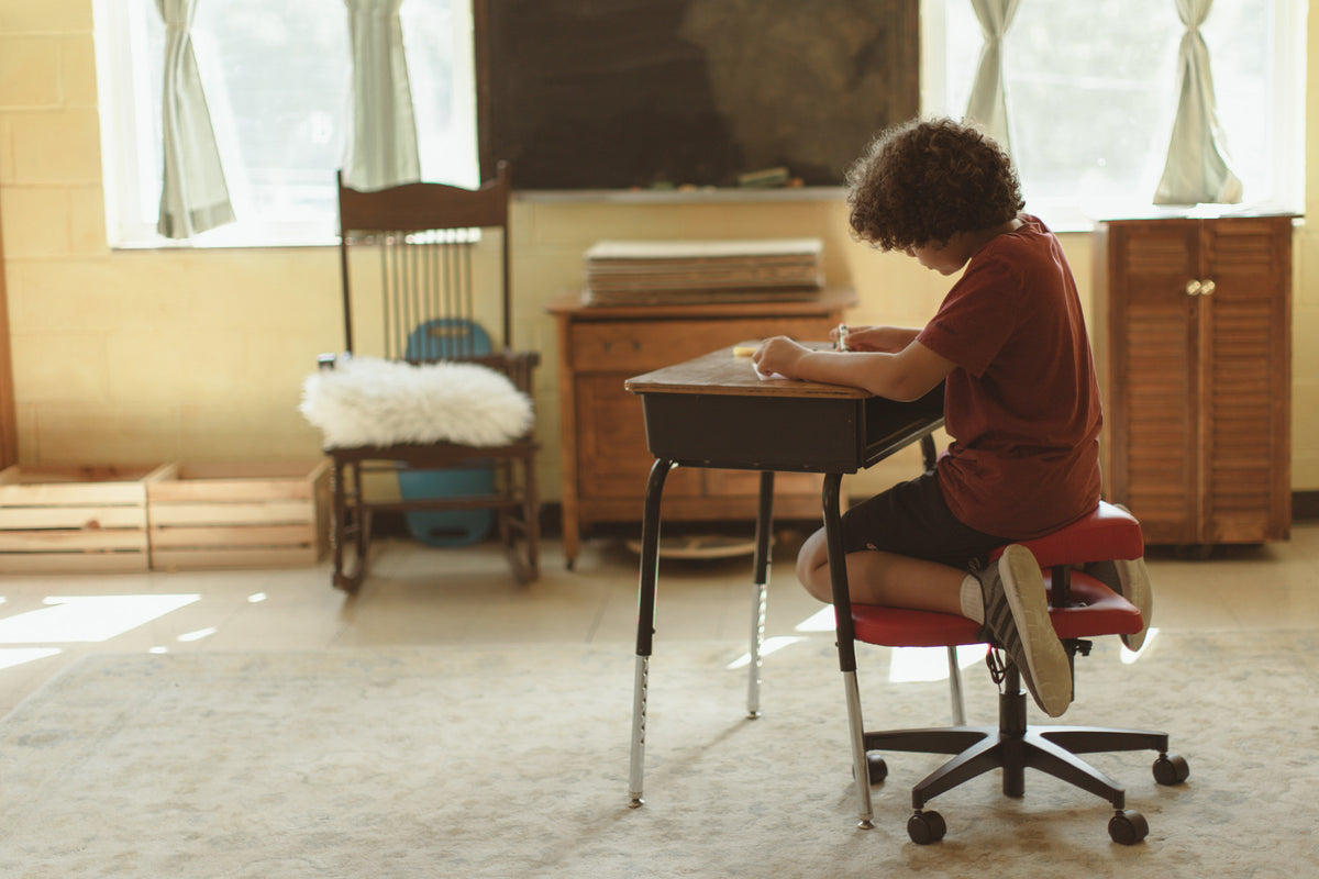 A child with curly hair sits on a fidget seat for kids at a small desk in a classroom, wearing a rust-red t-shirt and dark shorts while working on a piece of paper. The room has a chalkboard, wooden furniture, and warm natural light coming through the windows.