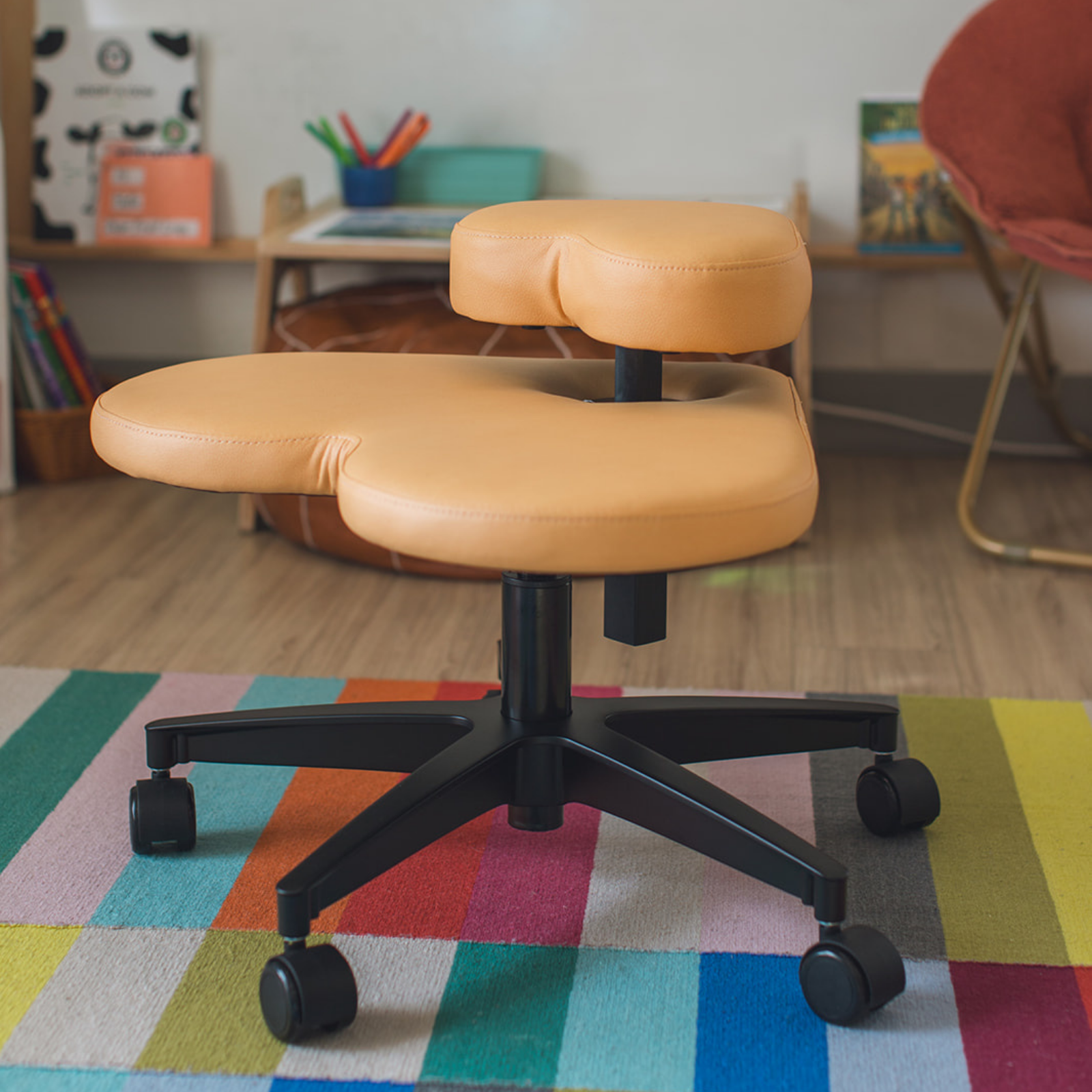 A fidget seat for kids - an ergonomic kneeling stool with a peach-colored contoured seat and a black five-star wheeled base, placed on a colorful striped rug in a home office setting.