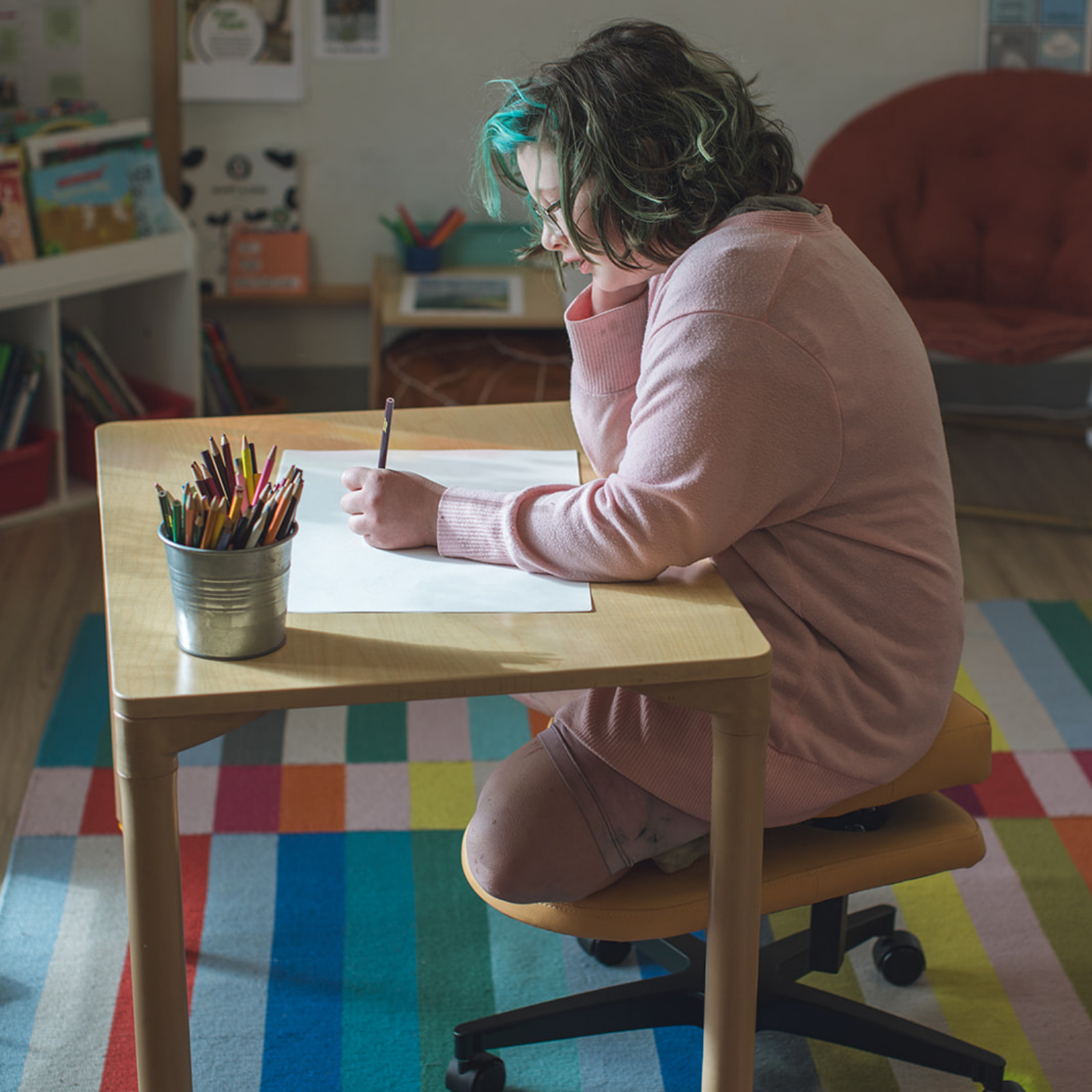 A kid with green-tinted hair sits on a fidget seat for kids at a small wooden desk, writing on a sheet of paper. A metal cup filled with colored pencils sits on the table next to them.