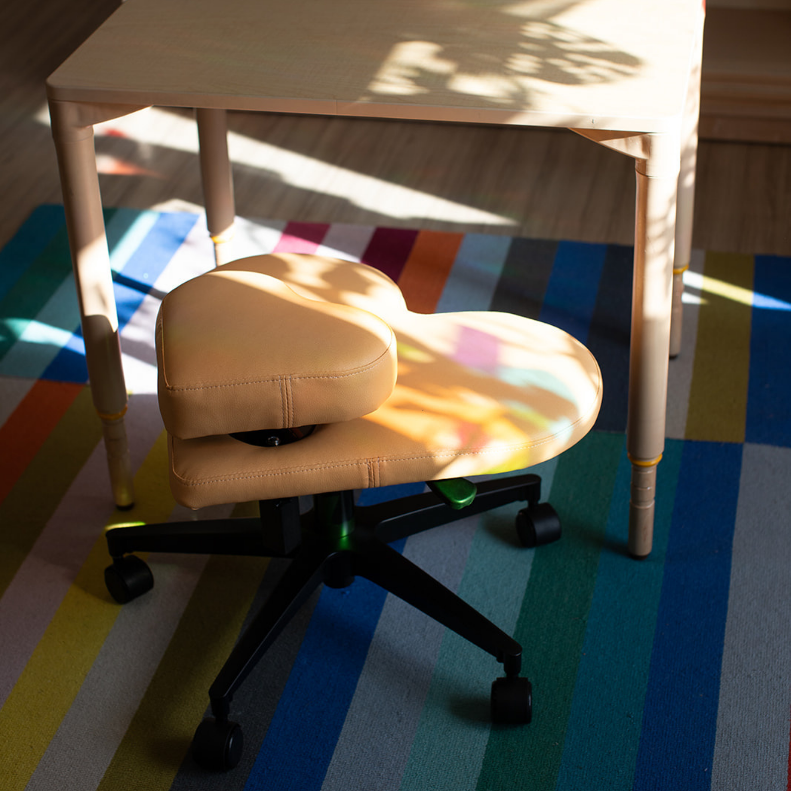 A fidget seat for kids in butterscotch yellow placed under a light wood table on a colorful striped rug with dappled sunlight.