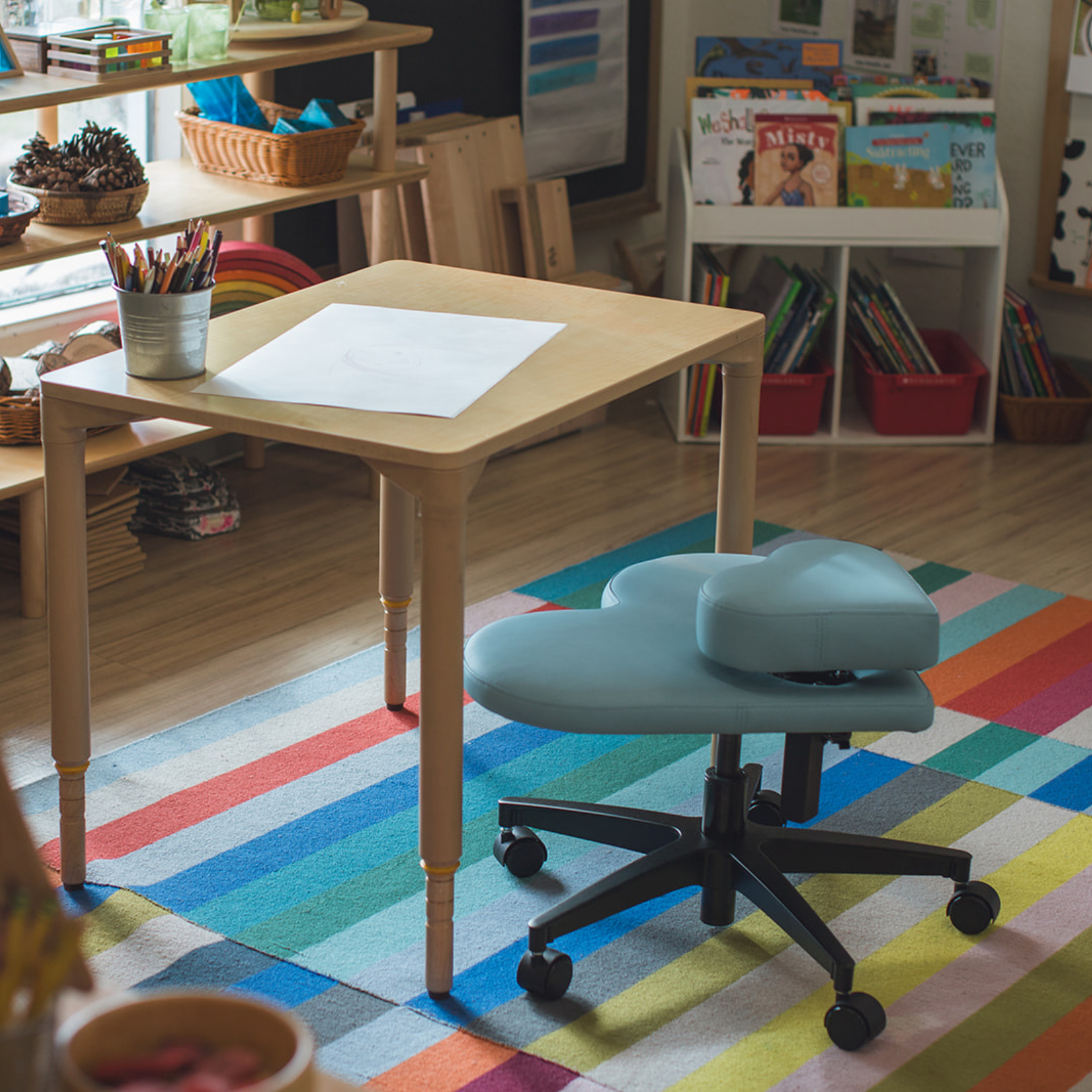 A light wood children's desk with a white drawing sheet on top, paired with a light blue fidget seat for kids on a colorful striped rug in a bright classroom setting.