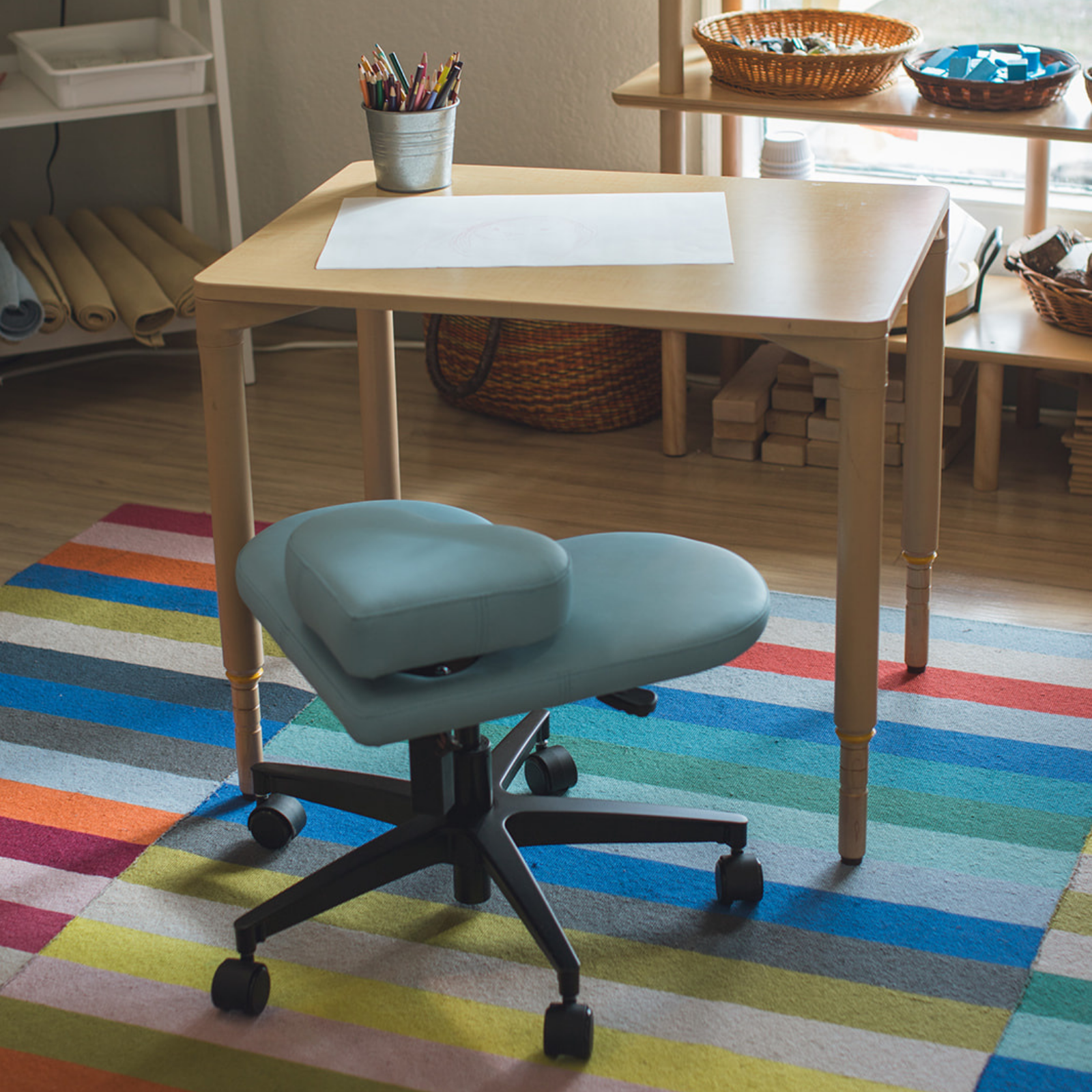 A light blue fidget seat for kids positioned in front of a wooden desk in a brightly lit room with a colorful striped rug.