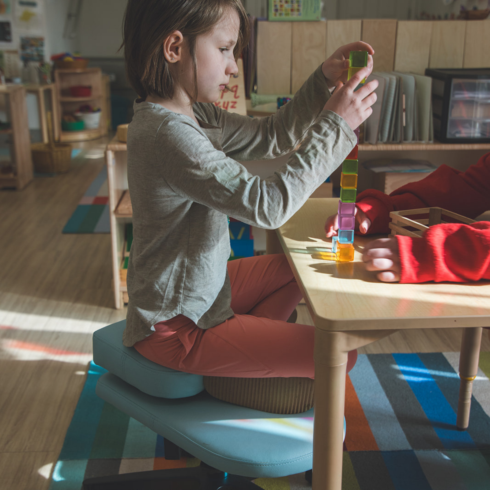 A child sits on a light blue fidget seat for kids at a small wooden table, wearing a gray long-sleeve shirt and rust-orange pants while stacking colorful blocks. The room has a warm, sunlit interior with wooden floors and shelves.