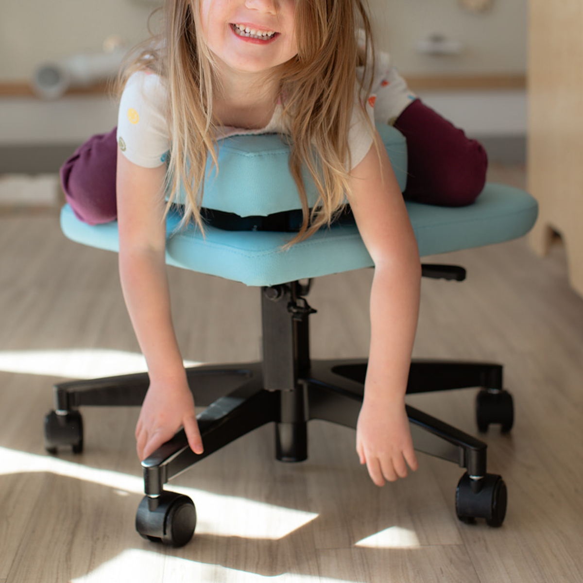 A young child smiling while leaning over a light blue upholstered fidget seat for kids with a black wheeled base in a sunlit indoor room.