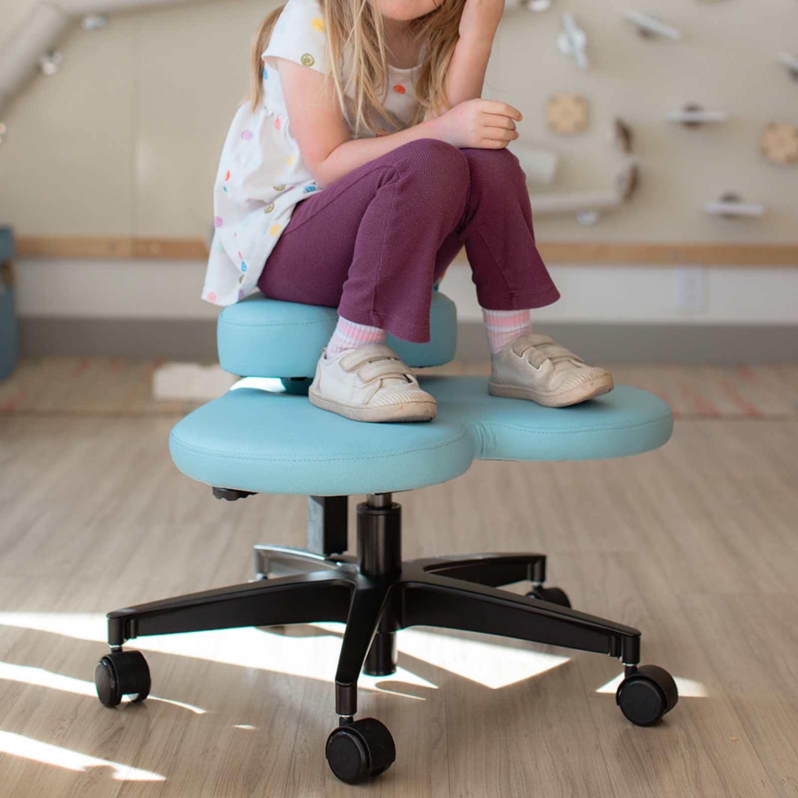 A child sits on a fidget seat for kids with a light blue cushion and a black wheeled base in a bright indoor room.