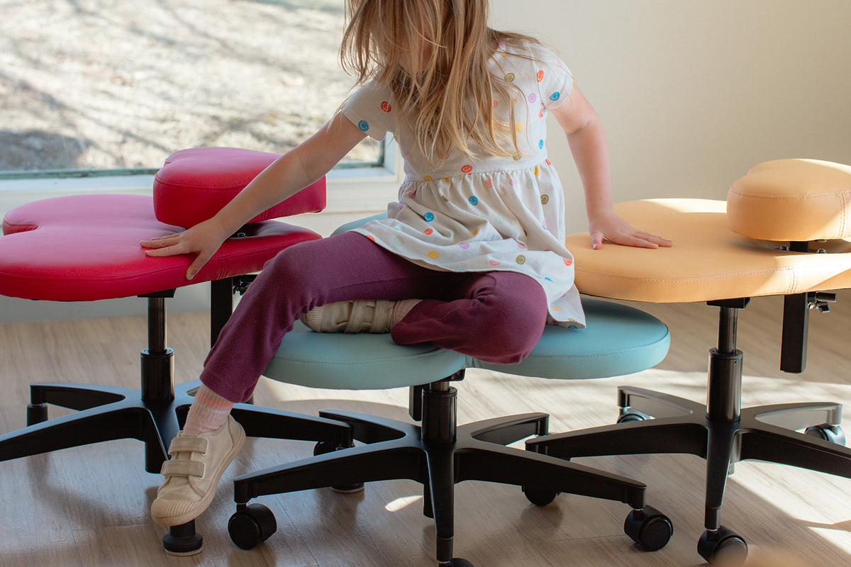 A young girl sits on a fidget seat for kids in cotton candy in a brightly lit room, with red and yellow versions shown beside her on black wheeled bases.