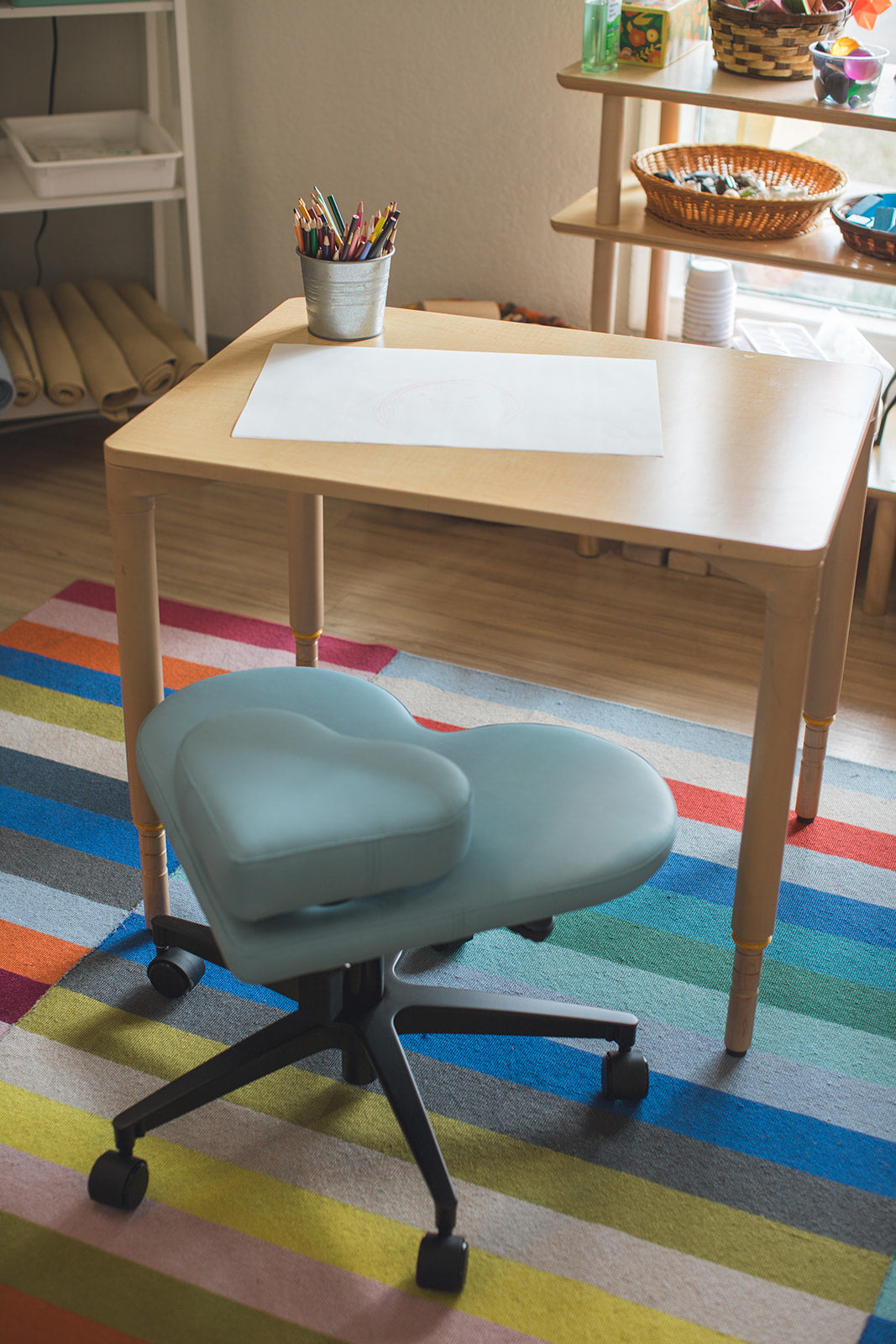 A light blue fidget seat for kids with heart-shaped cushions and black wheeled base, positioned in front of a light wood desk on a colorful striped rug.