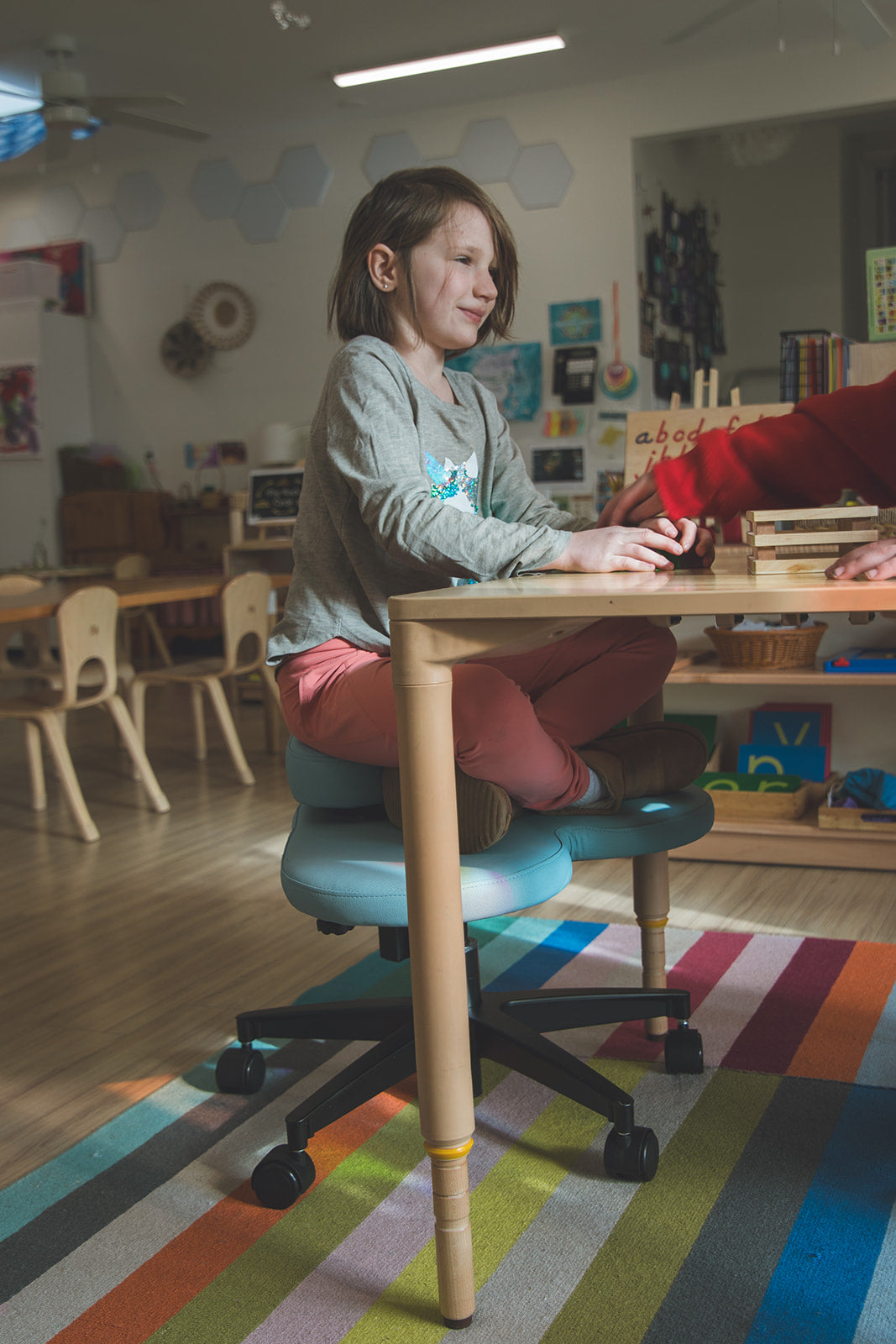 A child sits at a wooden table in a classroom, wearing a gray sweatshirt, pink pants, and a light blue Fidget Seat for kids with a black wheeled base. A colorful striped rug is visible on the floor.