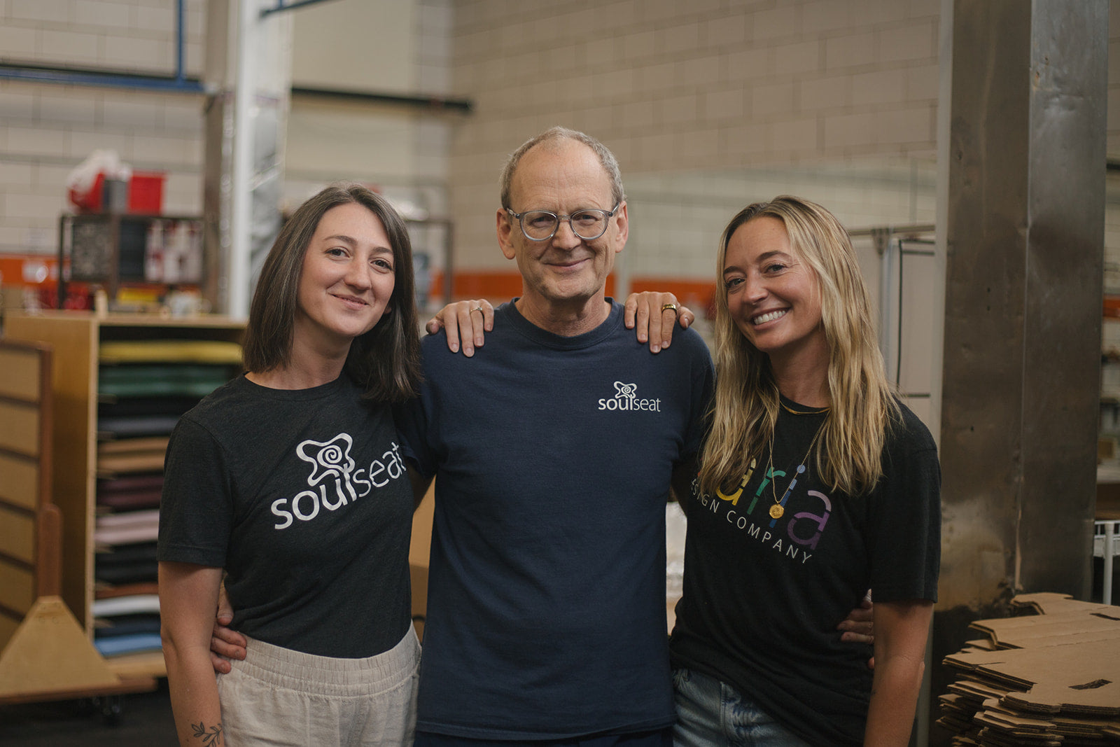 Smiling father with his two adult daughters wearing Soul Seat and Ikaria Design t-shirts in their factory